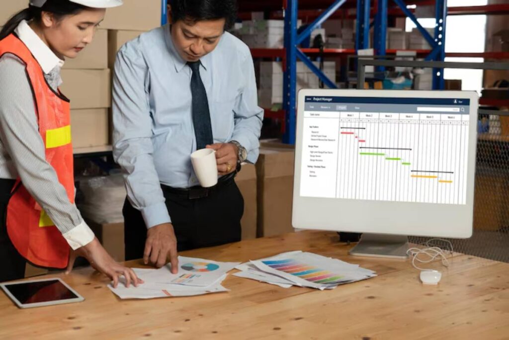 Two professionals review project plans on a desk with printed materials and a computer displaying a Gantt chart in a warehouse setting.