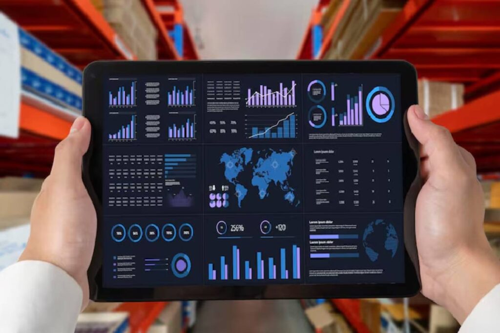 A person holds a tablet displaying various data visualizations and graphs in a warehouse setting. Bright colors against a dark background.
