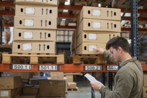 A warehouse worker inspects stacked cardboard boxes with labels, referencing a list while standing beside wooden pallets.