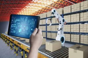 A hand holds a tablet displaying data while a robotic arm operates in a warehouse filled with stacked boxes.