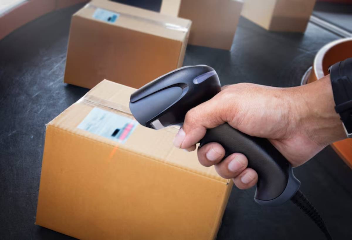 A hand holds a barcode scanner aimed at a cardboard box, with other boxes in the background, indicating inventory management.